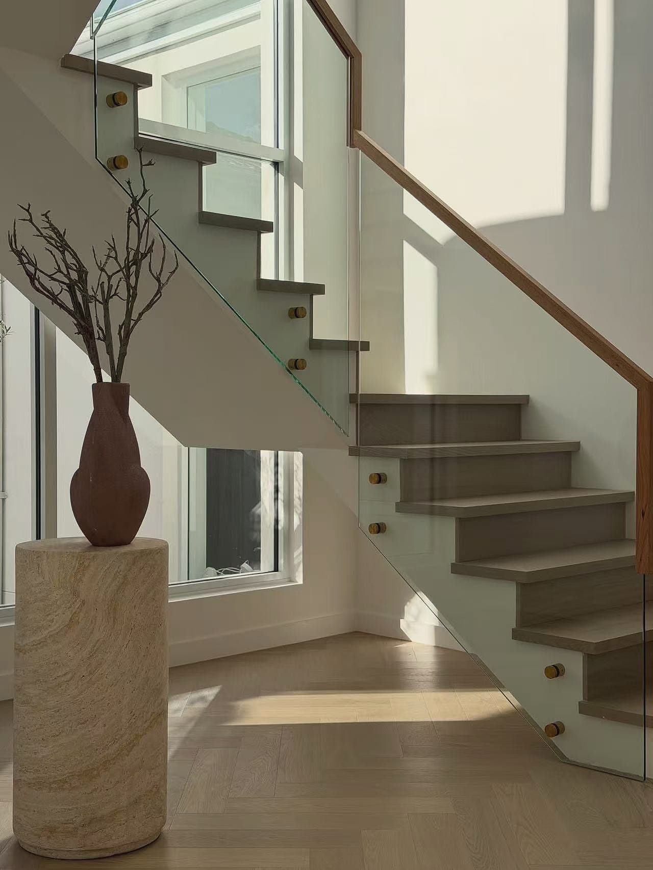 Timber stair with a glass balustrade rising above a herringbone floor, a vase on a stone plinth alongside