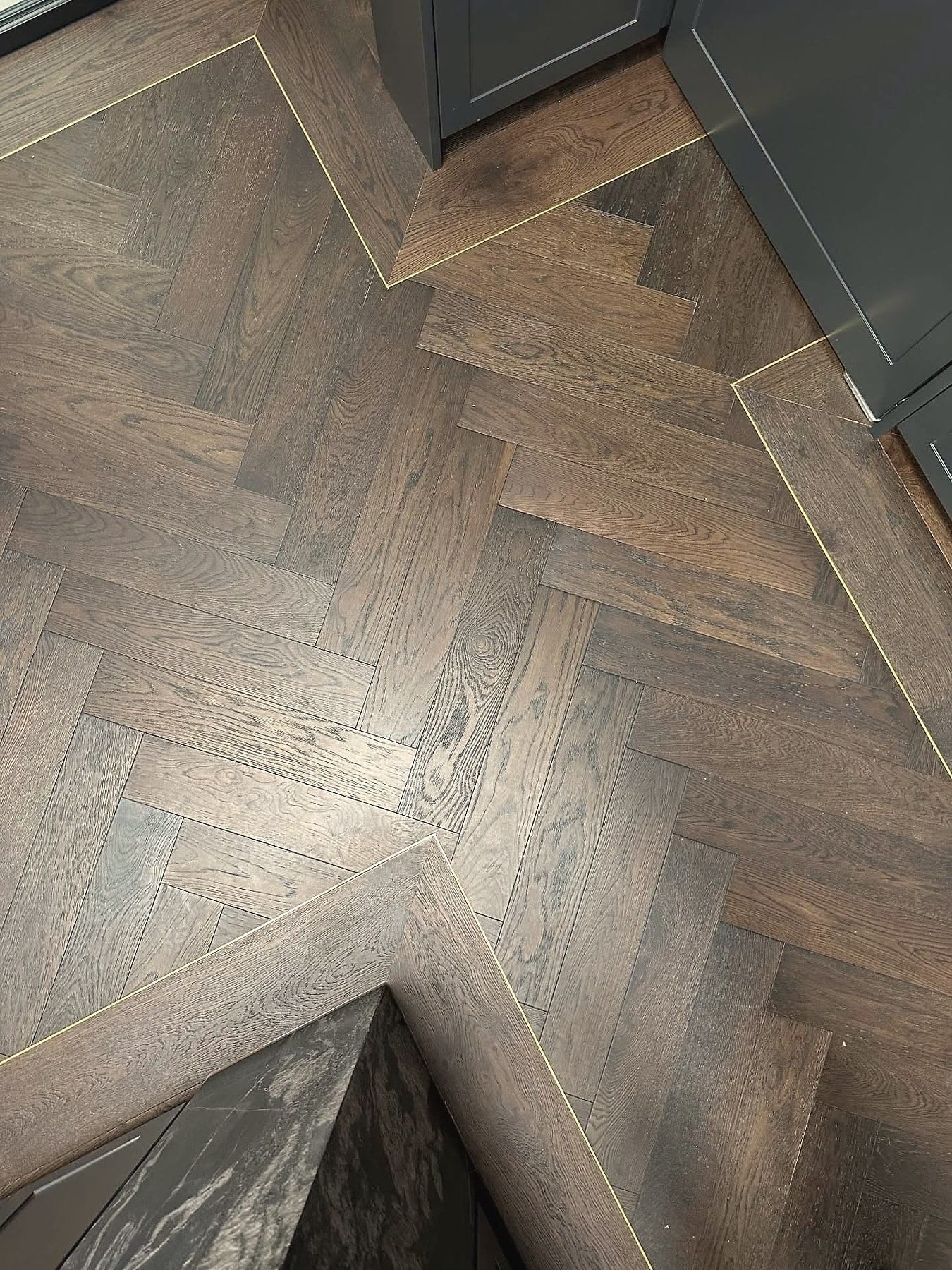 Dark smoked oak herringbone with a slim brass inlay strip and a mitred border, laid around dark joinery