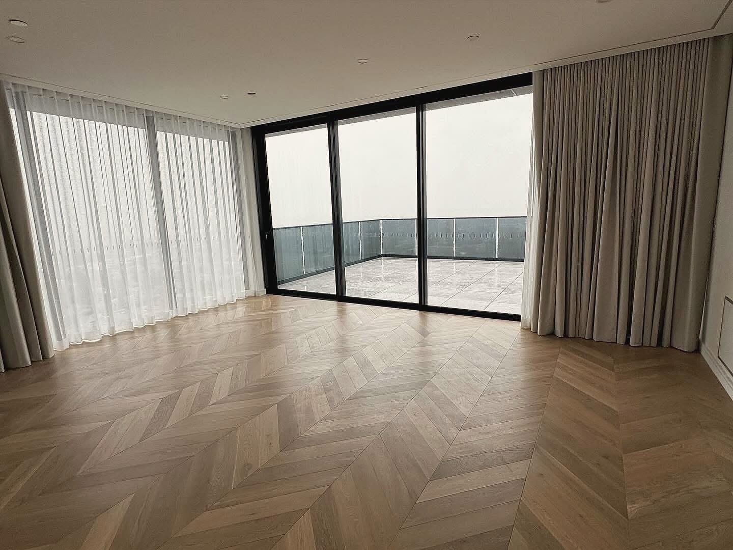 Empty room with pale chevron flooring, floor-to-ceiling glazing and full-length curtains